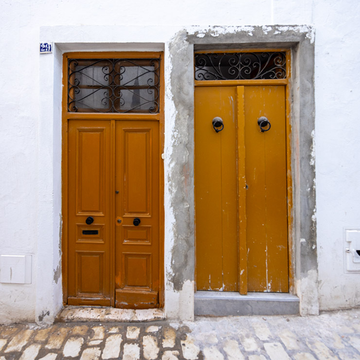 Two dark orange doors side by side in the Medina of Tunis, one paneled with a mail slot and the other plain with traditional knockers, set into a whitewashed wall.