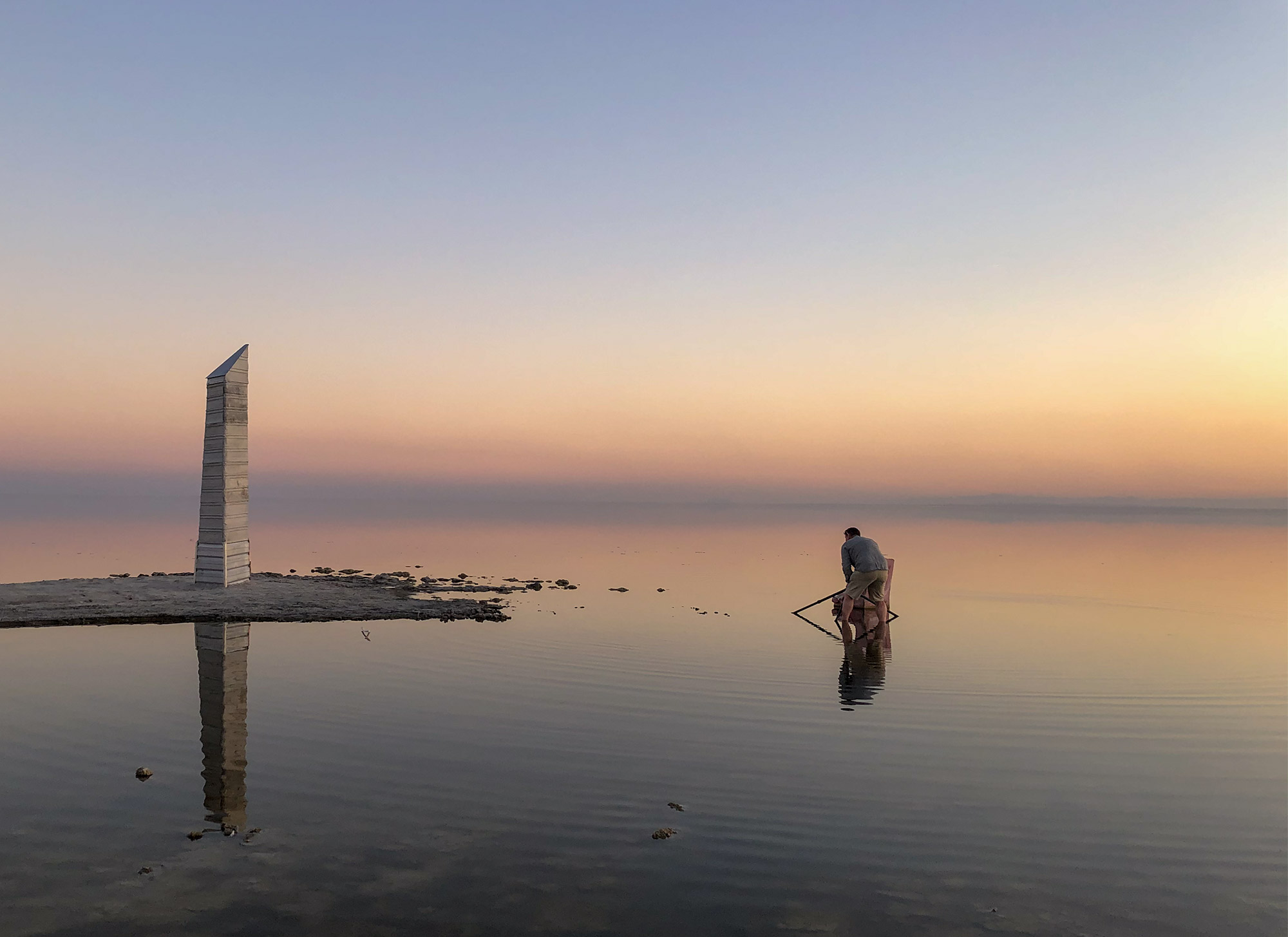 Erik Gauger stands in shallow water at the Salton Sea during sunset, adjusting a camera on a tripod. A tall, angular sculpture stands on a narrow sandbar to the left, with calm water and a pastel-colored sky in the background.