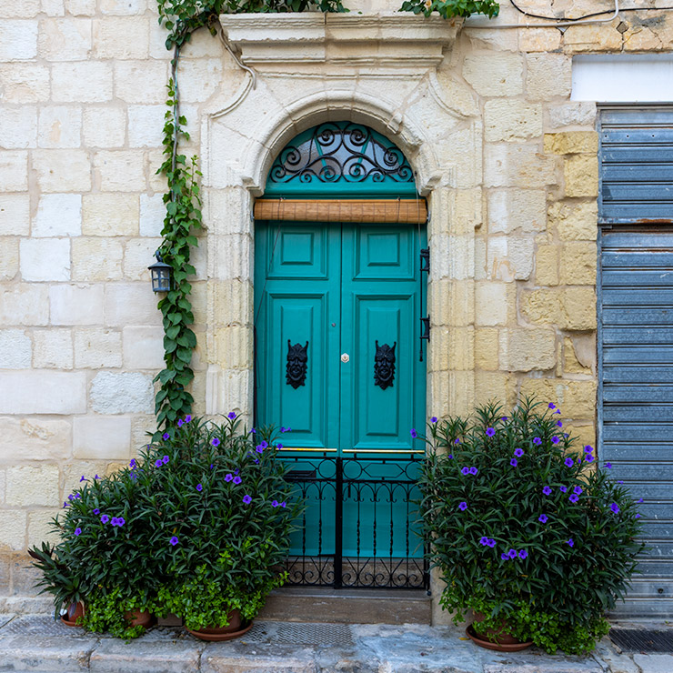 Green double door in Valletta framed by stone arch and potted purple flowers.