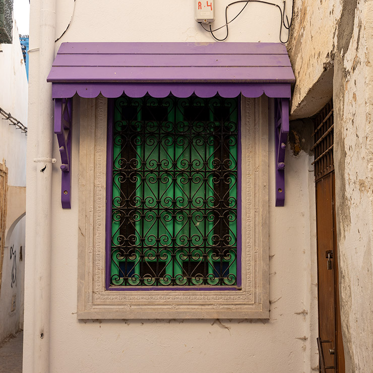 Green-shuttered window in the Medina of Tunis framed by ornate white stonework and protected by intricate black wrought iron bars, topped with a whimsical scalloped purple wooden awning and purple support brackets.