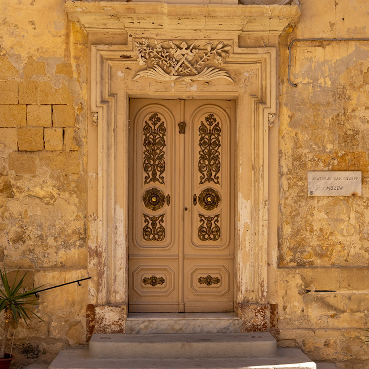Ornate double wooden door in honey-beige tones in Victory Square, Malta, with carved floral iron panels, circular brass medallions, and a decorative stone arch featuring symbolic religious carvings, set in a weathered limestone façade.