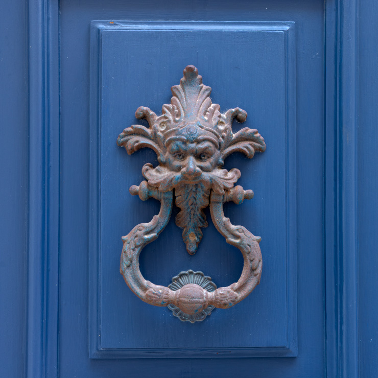Ornate cast iron door knocker on a deep blue wooden door in Senglea, Malta, featuring a stylized Green Man face with flowing leaf-like hair and beard, flanked by curled decorative elements and a scallop-shaped strike plate.