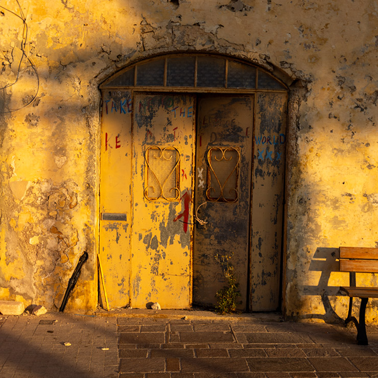 Weathered yellow door ajar in Marsaxlokk, with graffiti, iron grilles, and golden evening light.