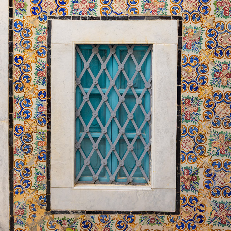 Turquoise blue window in the Medina of Tunis framed by white stone and protected by a diamond-patterned metal grille, surrounded by intricate, hand-painted ceramic tiles featuring floral motifs, golden arabesques, and cobalt blue scrollwork.