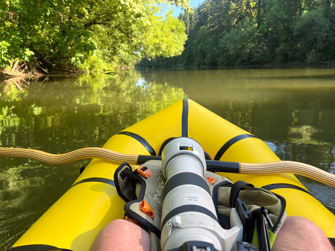 View from Erik Gauger's yellow Kokopelli packraft as he photographs wildlife on a calm, tree-lined river using a Canon 100–500mm lens. His legs and camera gear are visible in the foreground.