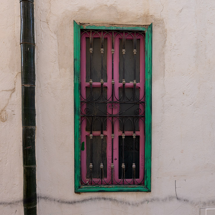 Tall narrow window in the Medina of Tunis with black wrought iron bars, painted pink on the inner frame and dark green on the outer frame, set in a weathered white stucco wall next to a vertical drainage pipe.