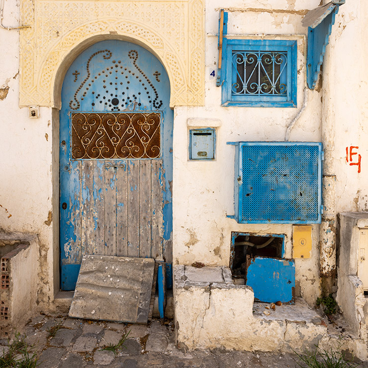 Weathered blue door and small window in Sidi Bou Said, Tunisia, set in a whitewashed wall beneath a carved cream archway, with iron grilles, flaking paint, utility boxes, and a worn stone threshold.