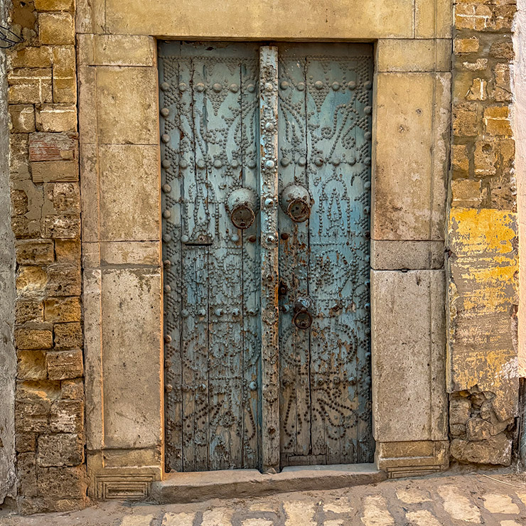 A weathered sky-blue metal-studded door with ornate geometric and floral patterns, set in a stone frame in the Medina of Tunis.