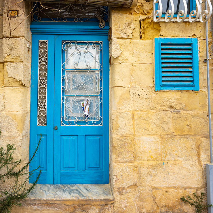 Sky blue wooden door in Valletta, Malta, set in a warm limestone facade, with ornate white wrought iron grilles over glass panels, a small vertical decorative inset, and a matching blue shuttered window nearby.