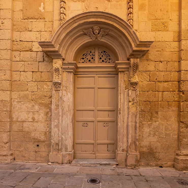 Tall beige door in Birgu, Malta, framed by an arched limestone portal with carved pilasters, a decorative fanlight grille, and a sculpted angel keystone above the arch, set into a weathered stone façade.
