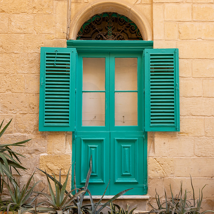 Teal-painted shuttered window in Birgu, Malta, with glass panes and raised wood paneling on the lower half, flanked by louvered shutters and topped by a rounded transom with ornate black ironwork, set in a warm limestone wall above a bed of aloe plants.