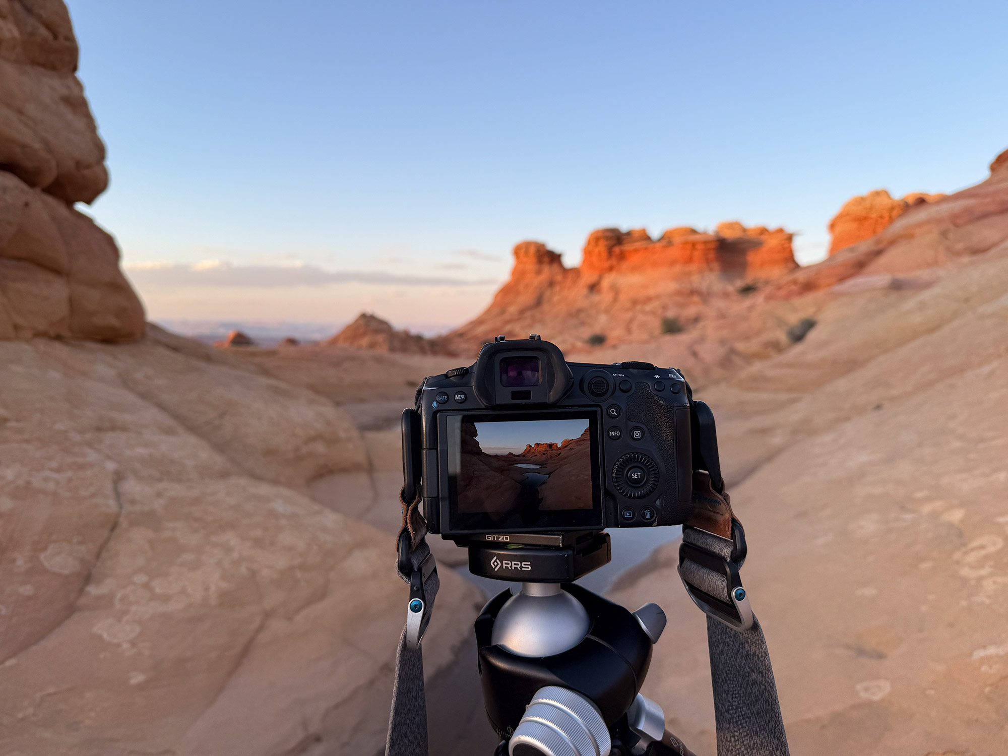 A Canon R5 camera on a tripod captures the red rock formations of the Paria Vermilion Cliffs Wilderness at sunset. The camera screen shows the landscape being photographed, surrounded by smooth sandstone terrain.