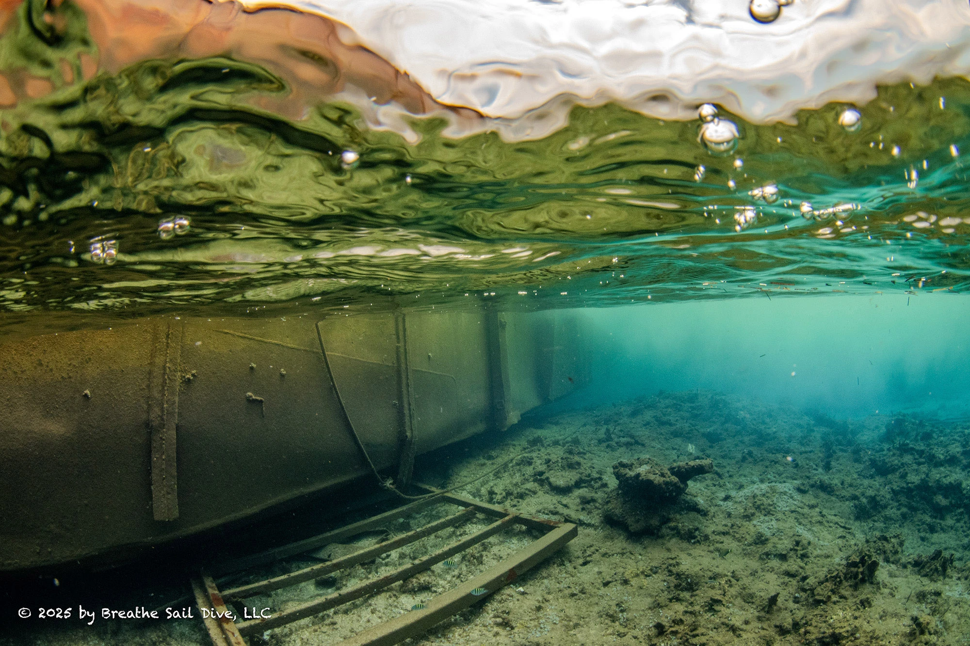 Underwater view of the grounded barge at Fowl Cays National Park, showing its rusted metal hull pressed against the seafloor. Coral rubble and damaged reef structure lie beneath it, with sediment clouding the water and small fish swimming near the wreckage. The image captures the destruction caused by the barge resting directly on the reef.