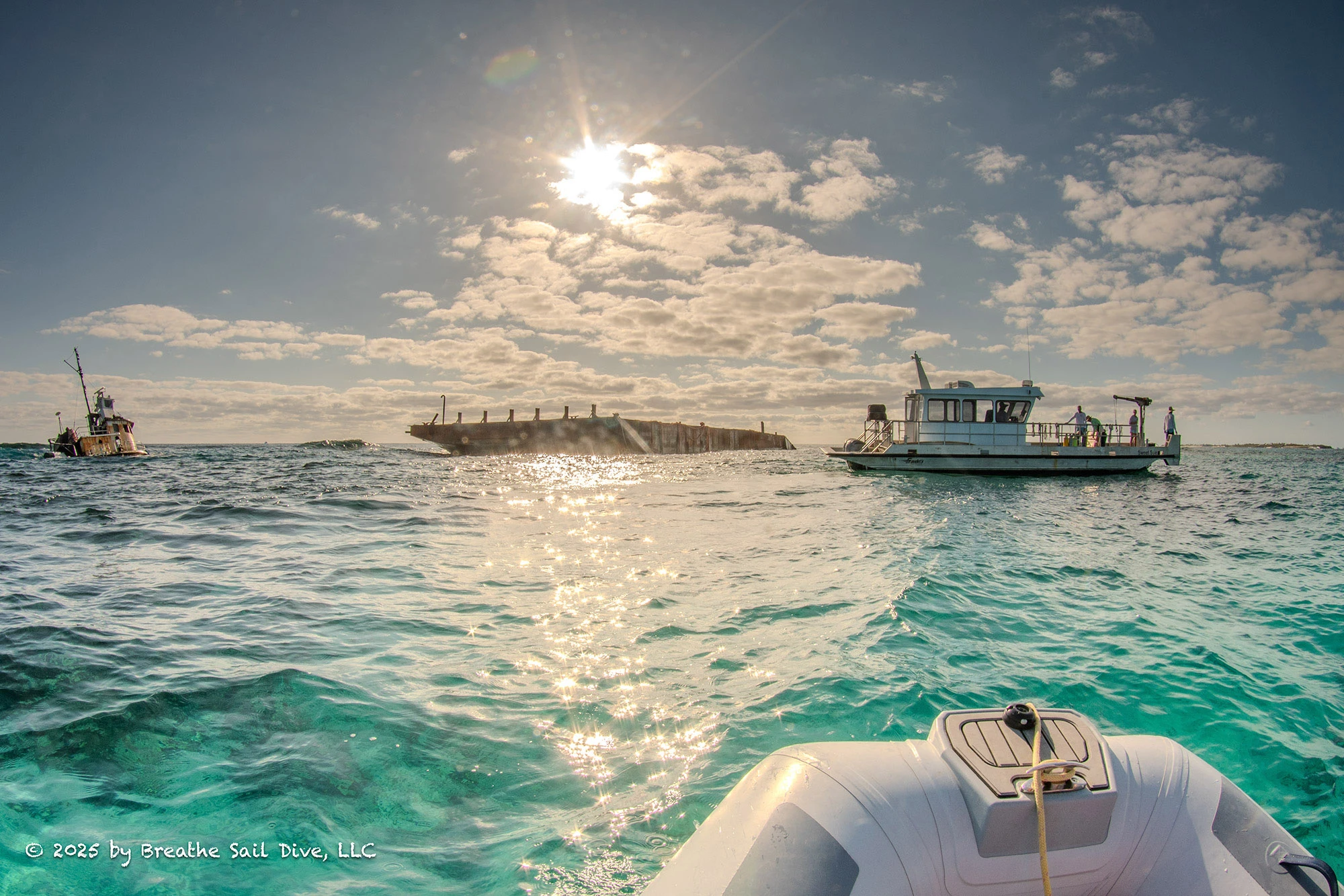 Volunteers aboard a small research vessel work near a large rusting barge and partially sunken tugboat stranded on the reef at Fowl Cays National Park. The turquoise water is calm under a bright sun, and a small inflatable boat floats in the foreground. The damage from the grounded vessels contrasts sharply with the clear, pristine marine environment.
