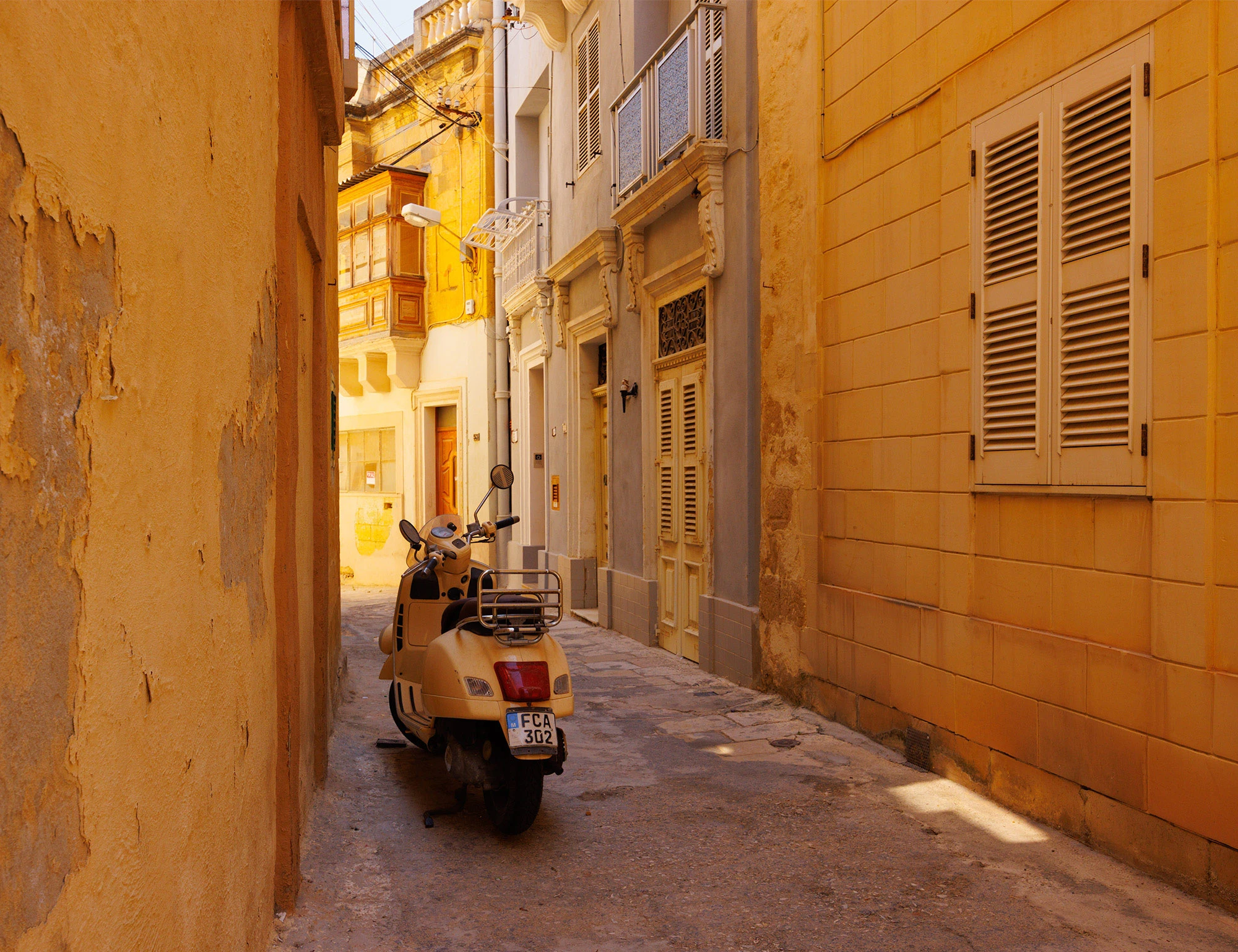 Sunlit alley in Birgu, Malta, flanked by honey-colored limestone buildings with shuttered windows, wooden balconies, and a parked cream Vespa.