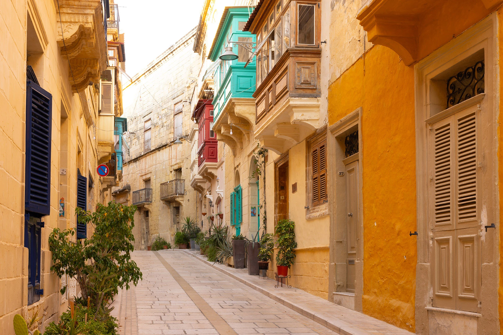 A sunlit alley in Birgu, Malta, lined with colorful balconies, shuttered doors, and potted plants along honey-colored stone walls.