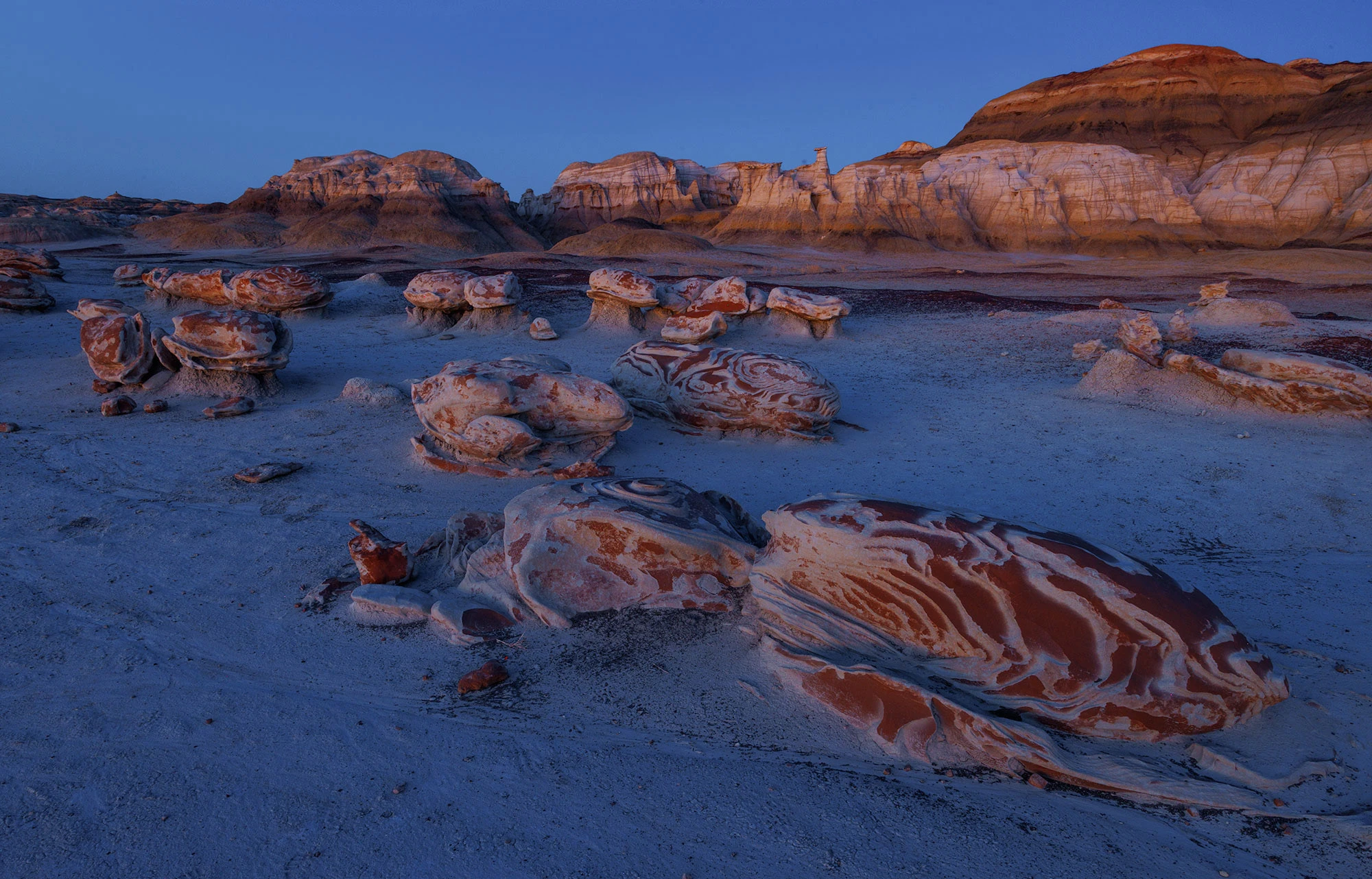 Swirled, pancake-like rock formations in the Bisti Badlands glow under twilight in the Bisti/De-Na-Zin Wilderness, with striped cliffs rising in the background.