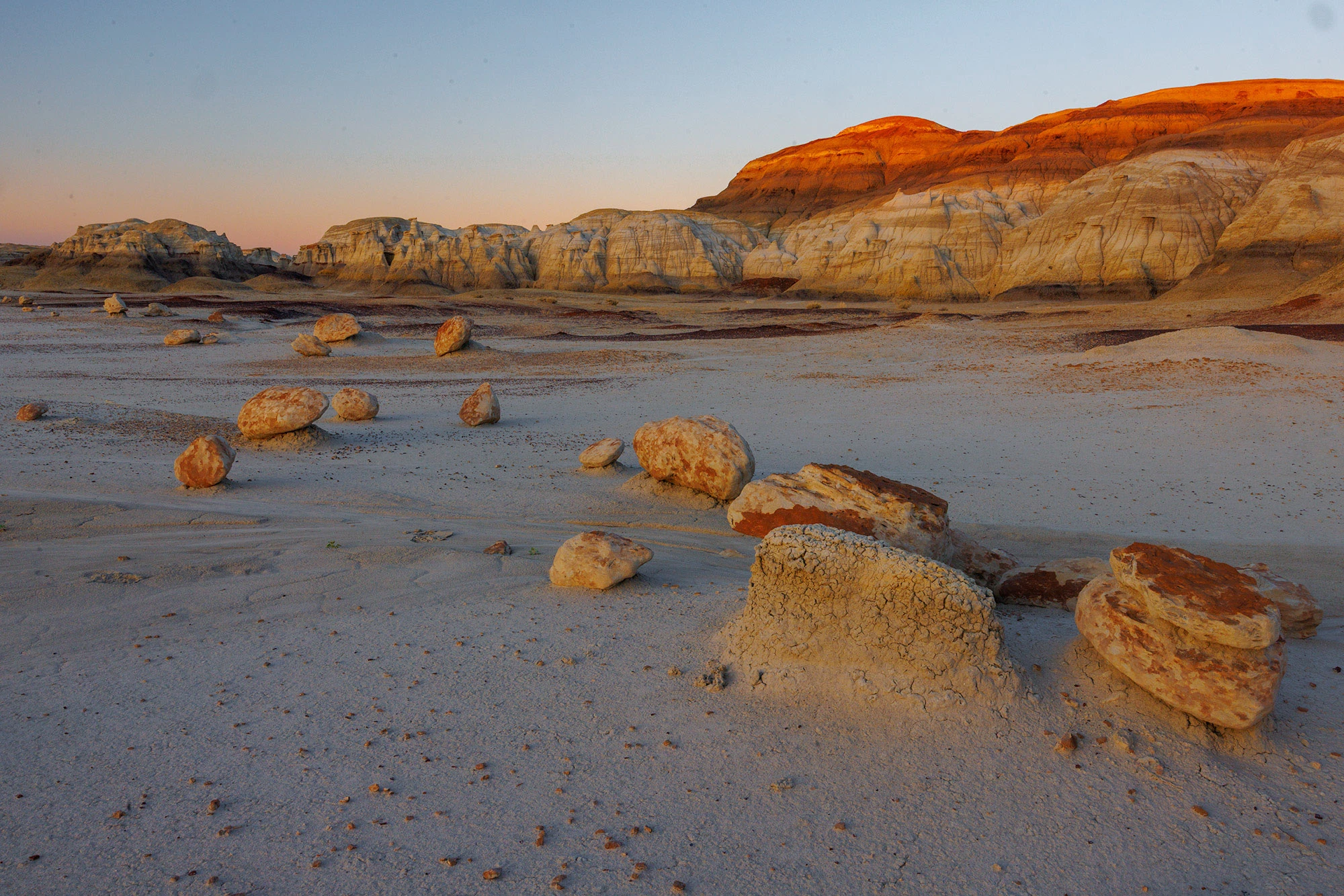 Evening light on round, eroded boulders scattered across a pale clay flat near striped badlands cliffs in the Bisti/De-Na-Zin Wilderness.