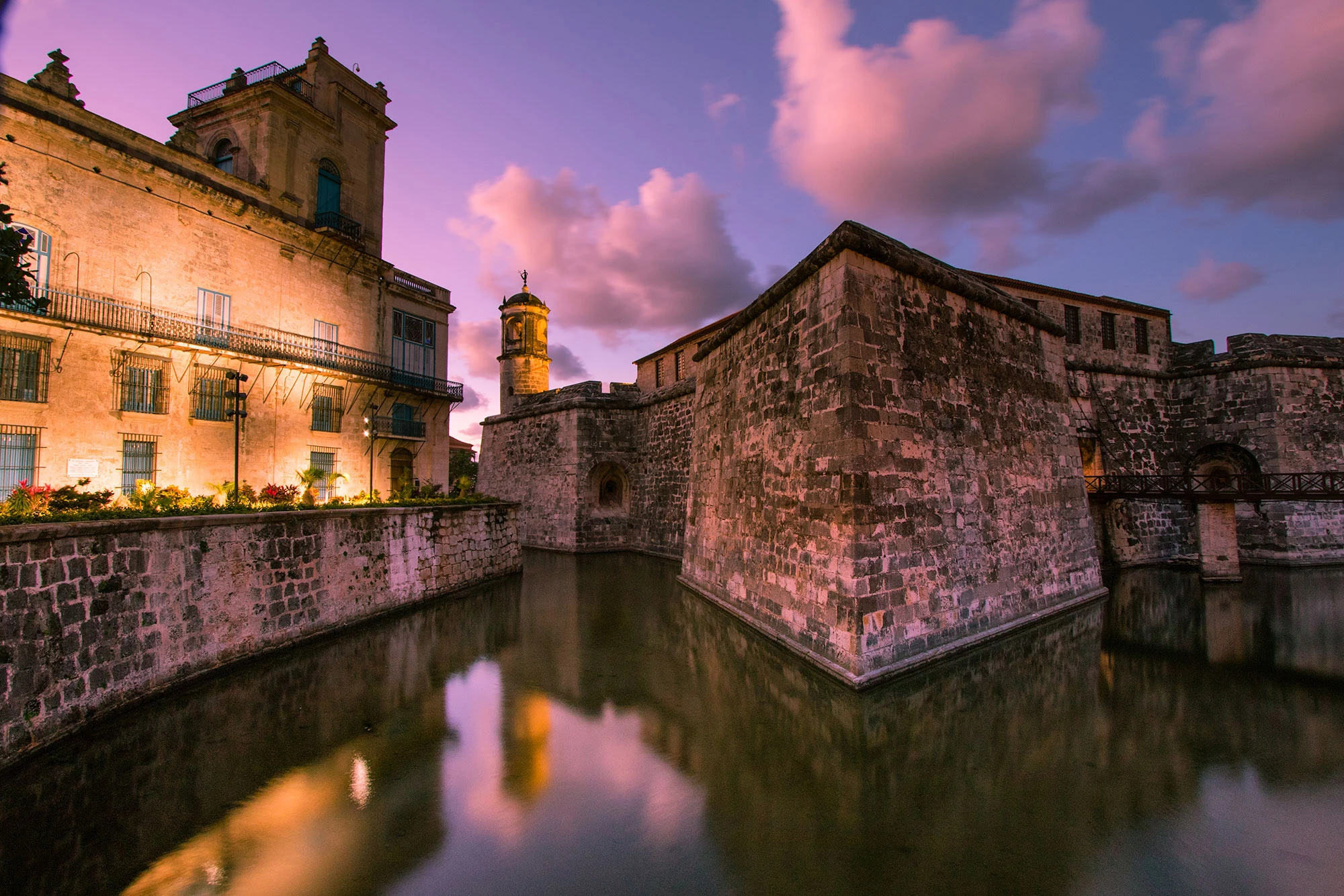 Castillo de la Real Fuerza at dusk in Habana Vieja: pink twilight clouds reflected in the moat, massive stone bastions and a small tower glowing under warm lights, with a narrow footbridge at the entrance.