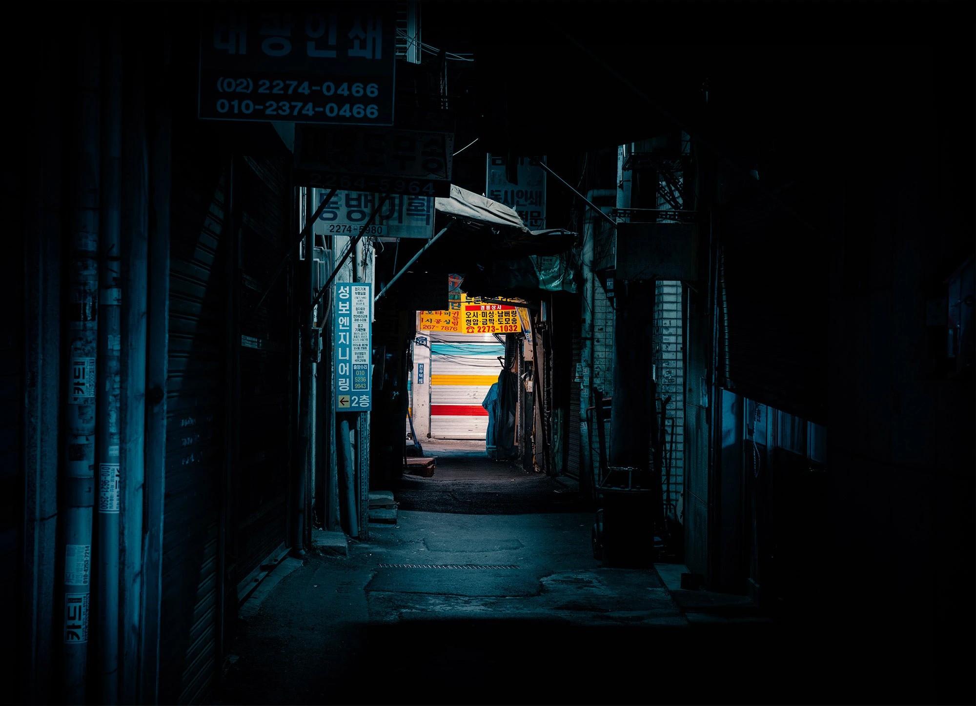 A narrow Seoul alley at night glows with overlapping signage, neon light, and cluttered facades. Korean characters light the path where pedestrians wander beneath dangling wires and the buzz of city life.