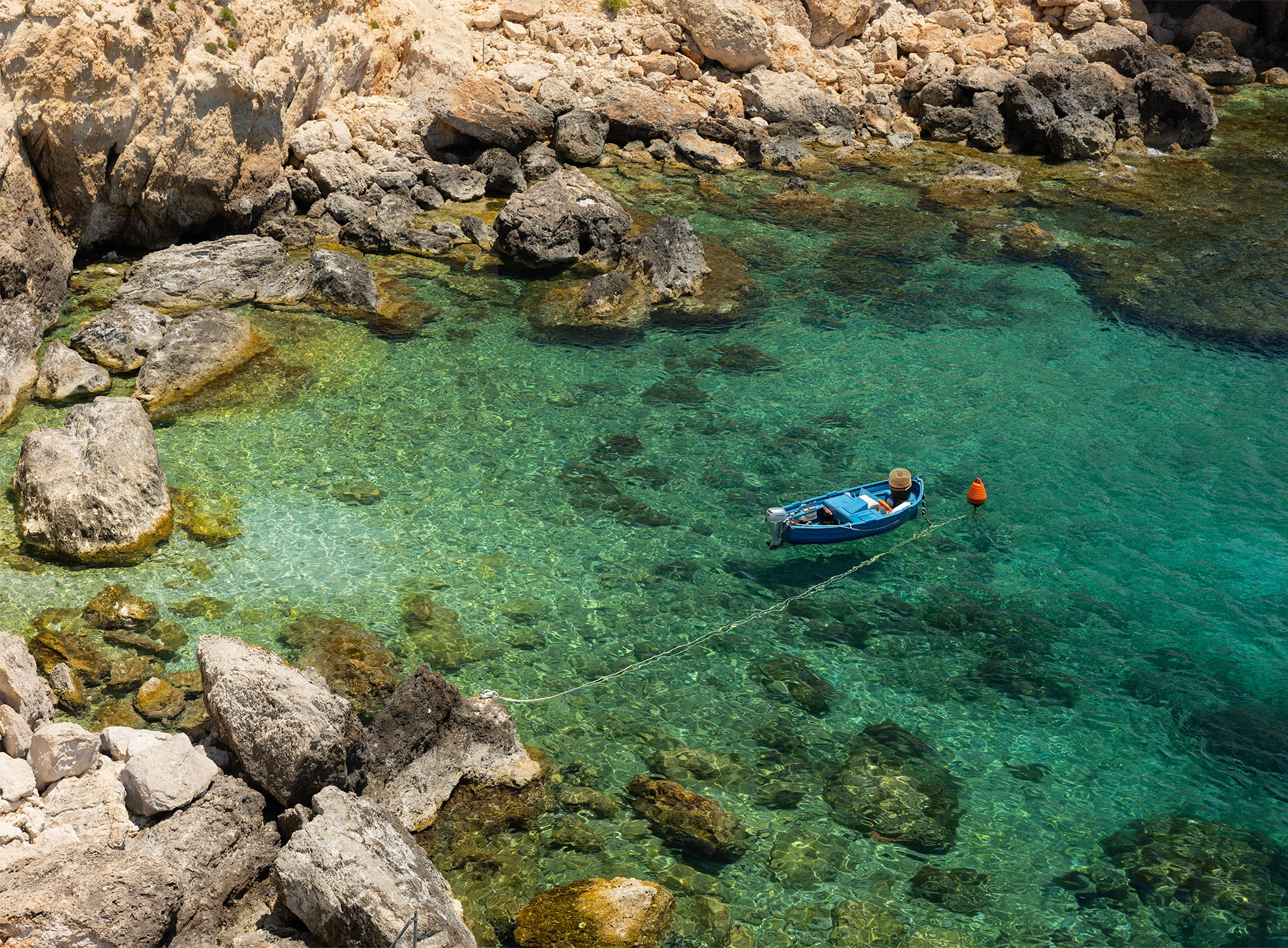 A small blue boat floats in the crystal-clear turquoise waters of Għar Lapsi, Malta, surrounded by rocky shoreline and submerged stones. The scene captures the serene beauty of this coastal swimming hole.