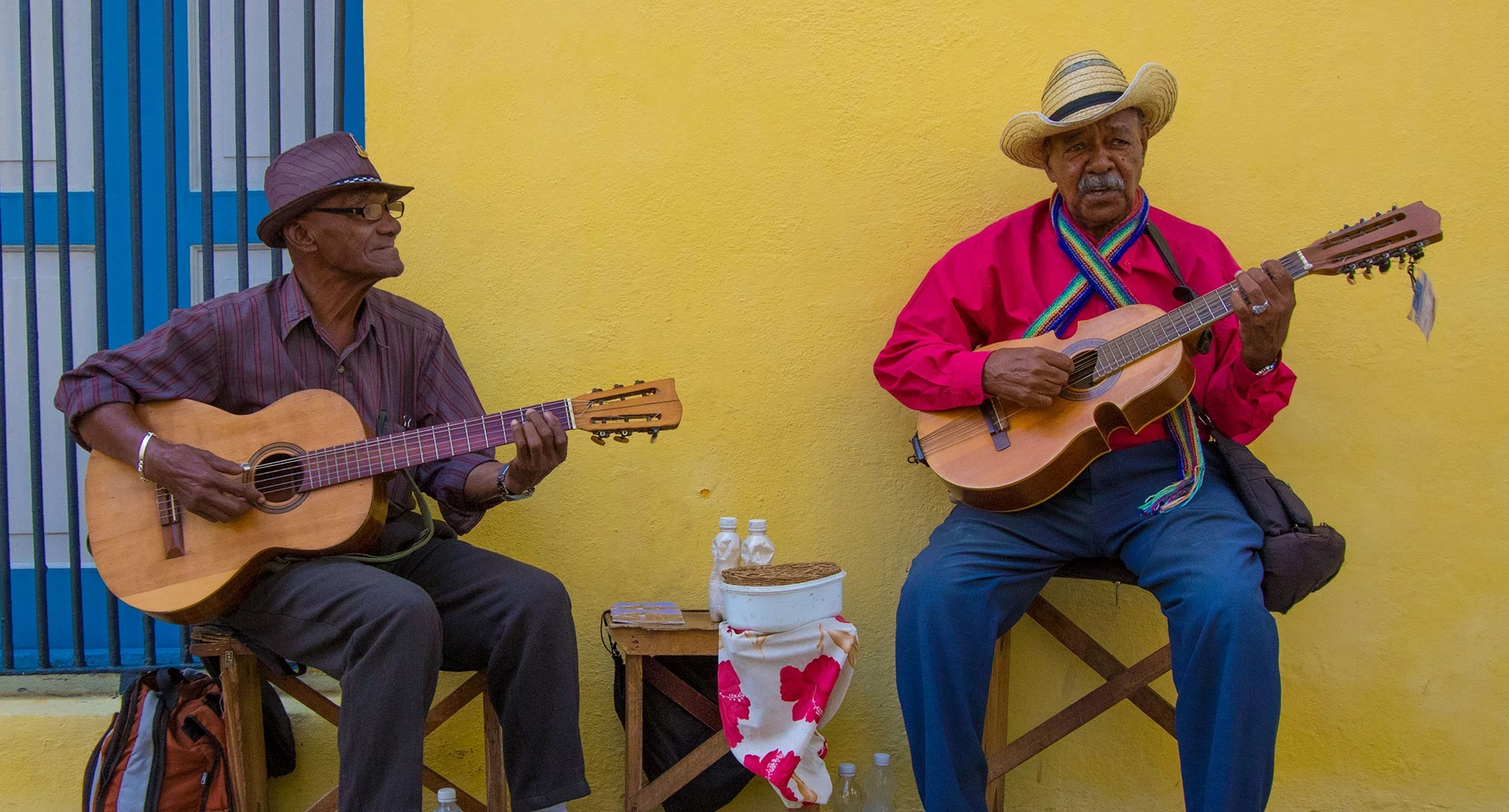 Street musicians in Habana Vieja: two Cuban guitarists seated against a bright yellow wall—one in a brown fedora, the other in a straw hat and bright red shirt with a rainbow scarf—playing acoustic guitars beside a blue-barred window.