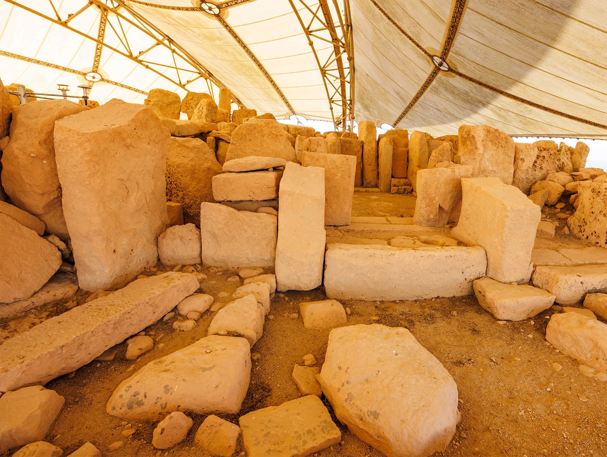 Interior view of Ħaġar Qim megalithic temple in Malta, where massive honey-colored limestone slabs rise beneath a white protective canopy. The stones are weathered and sunlit, evoking Malta's Neolithic past.