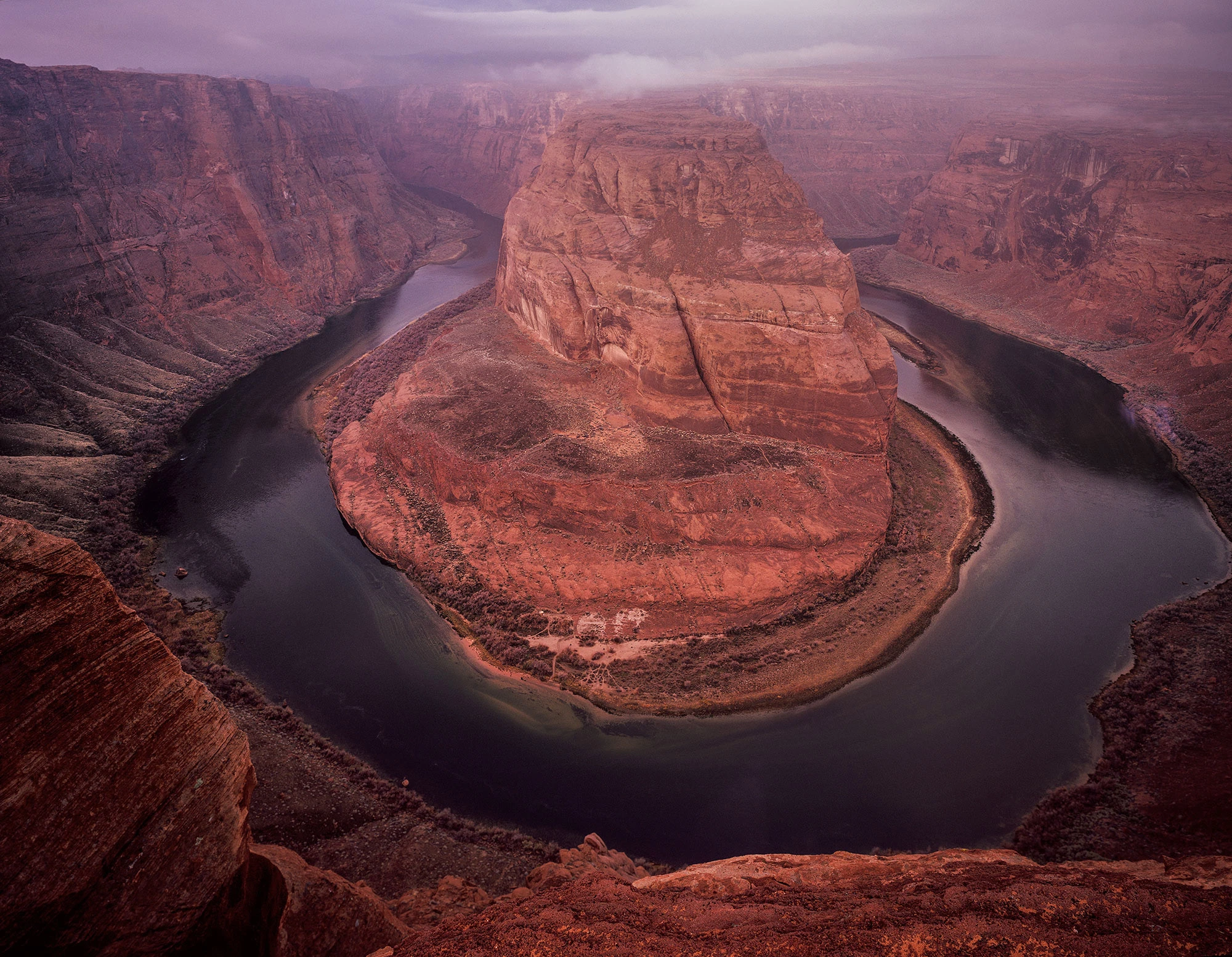 Predawn view of Horseshoe Bend on the Colorado River, captured under soft purple light with low-hanging clouds clinging to the canyon rim. The river forms a perfect oxbow around the massive sandstone butte, its waters dark and glassy in the early light. Taken en route to Coyote Buttes, this image marks the beginning of a winter journey into the Vermilion Cliffs wilderness.