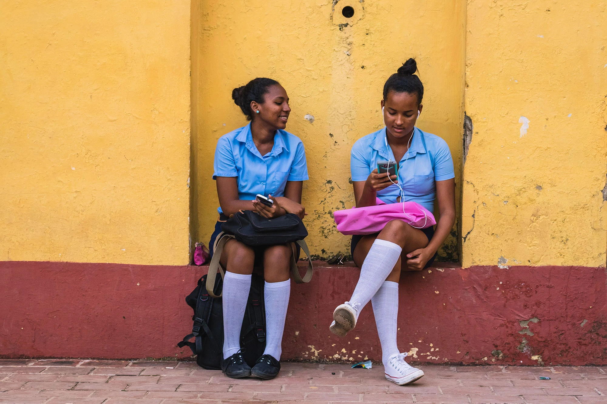 Two Cuban schoolgirls in blue uniforms sit against a sun-faded yellow wall in Trinidad, sharing music on their phones. One smiles while holding her bag, the other scrolls through her screen with earphones in. Their white knee socks and the cracked, red-painted curb capture a moment of modern youth framed by the texture of colonial Cuba.