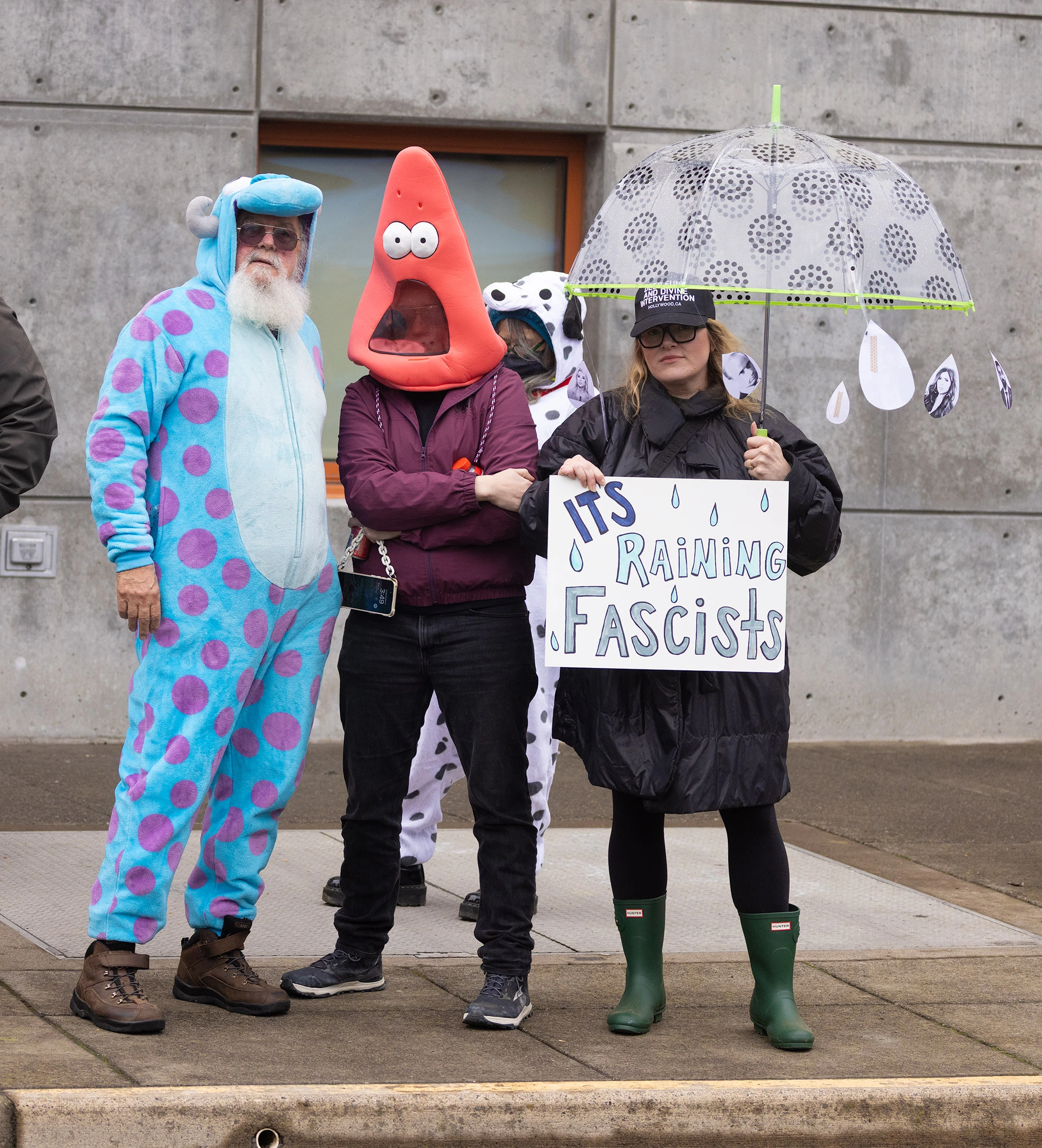 Protesters outside the Portland ICE building wear playful costumes, including a blue monster suit and a red cartoon headpiece, while one person holds an umbrella decorated with cutouts and a sign that reads 'It's Raining Fascists.'