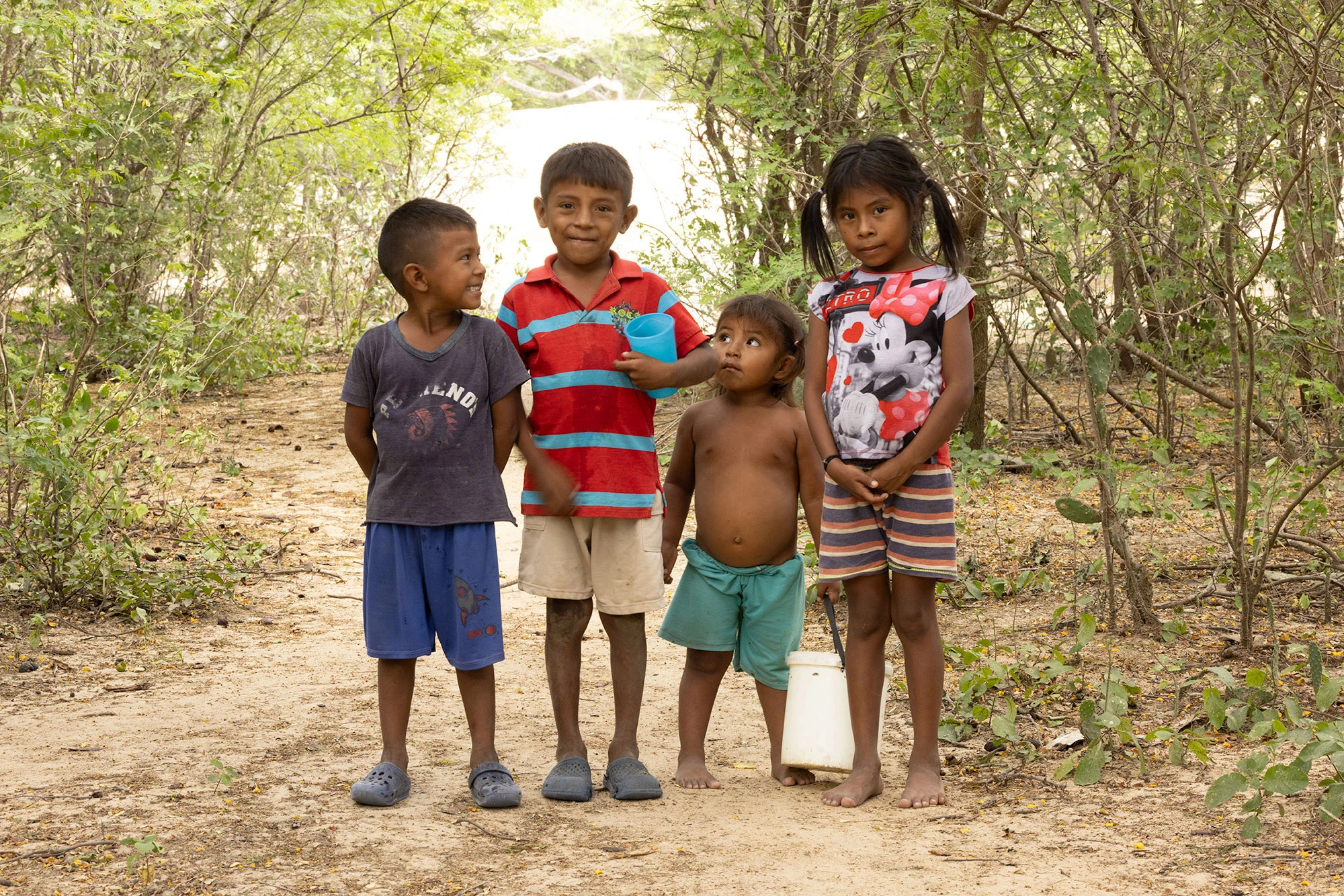 Wayuu children play in the dusty scrub forest of La Guajira, Colombia, surrounded by thorn trees, shadows, and the quiet resilience of their ancestral lands. A breeze lifts the dust as the light filters through dry canopy above.