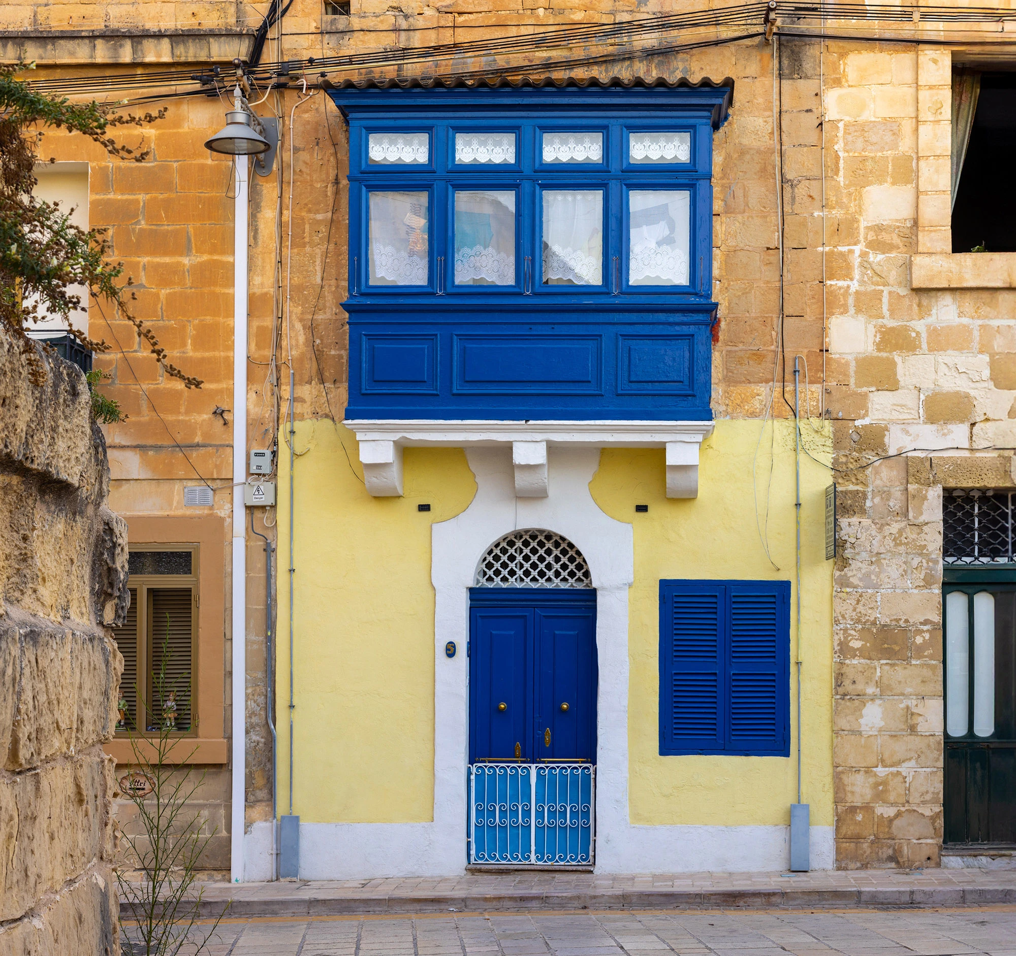 A traditional Maltese townhouse in Mdina, its limestone walls painted a soft yellow and framed by weathered golden stone. A deep cobalt blue door with brass handles sits at the center, topped with a white arched frame and lattice window. Above it, a vivid enclosed wooden balcony—painted the same striking blue—juts outward, its lace curtains hinting at domestic life inside. A matching blue shuttered window and small iron gate echo the color, standing out against the muted stone. The scene captures Mdina's blend of Mediterranean warmth, North African influence in its arches, and a lived-in intimacy that persists even within the Silent City's walls.