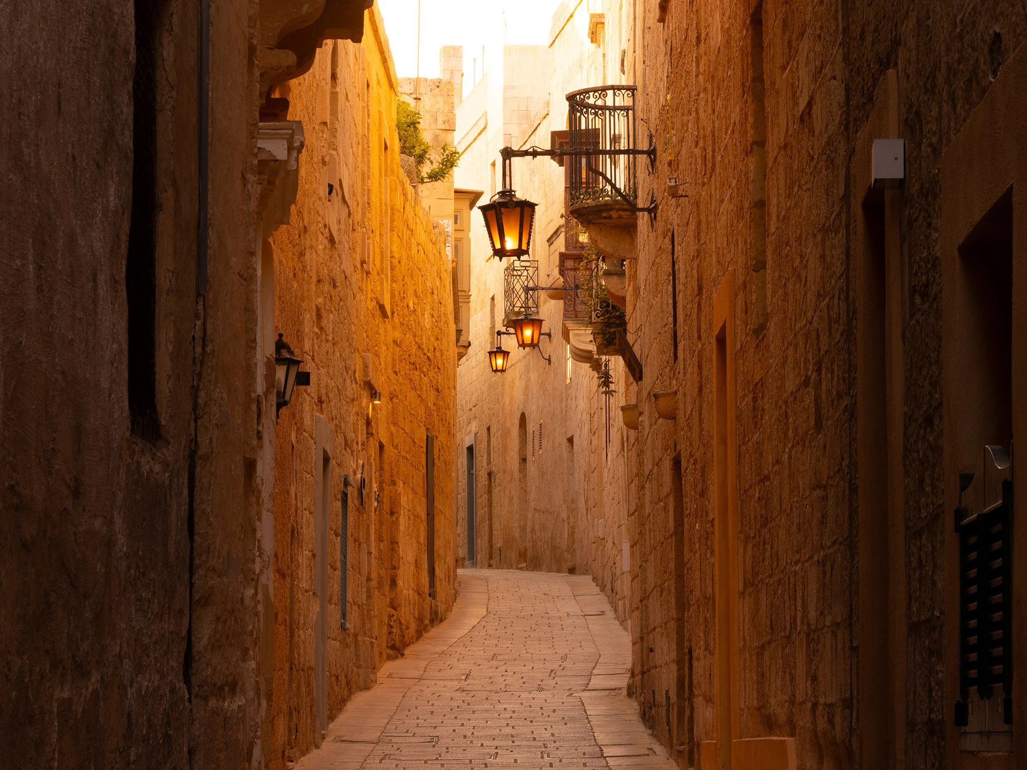 A narrow, winding alleyway in Mdina, Malta, its pale honey-colored limestone walls glowing with warm evening light. Iron balconies jut overhead, some with flower pots, while ornate black lanterns hang from the walls, casting amber light onto the cobbled street below. The alley rises gently uphill, curving out of sight, suggesting both secrecy and timelessness. This is the Silent City, once deliberately contracted under Arab rule, now preserved as a semi-rural enclave and tourist draw, where the stones themselves hold the memory of shifting dynasties and the cultural crosscurrents that shaped Malta.