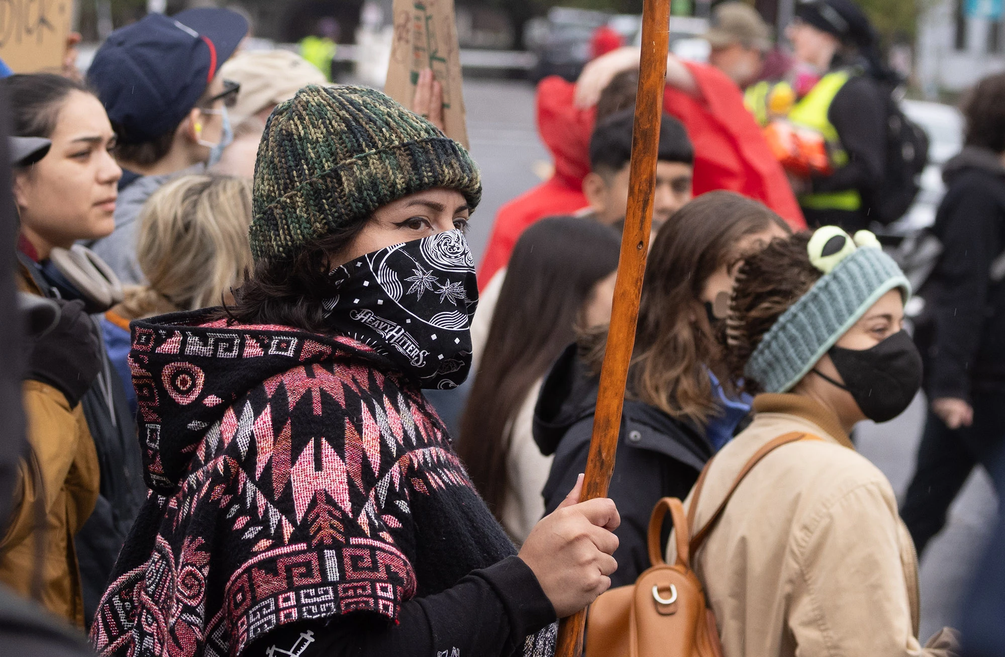 A masked protester wearing a patterned poncho and knit cap stands among the Portland crowd during an anti-ICE and anti-Trump demonstration, holding a wooden staff and looking resolutely toward the federal building.