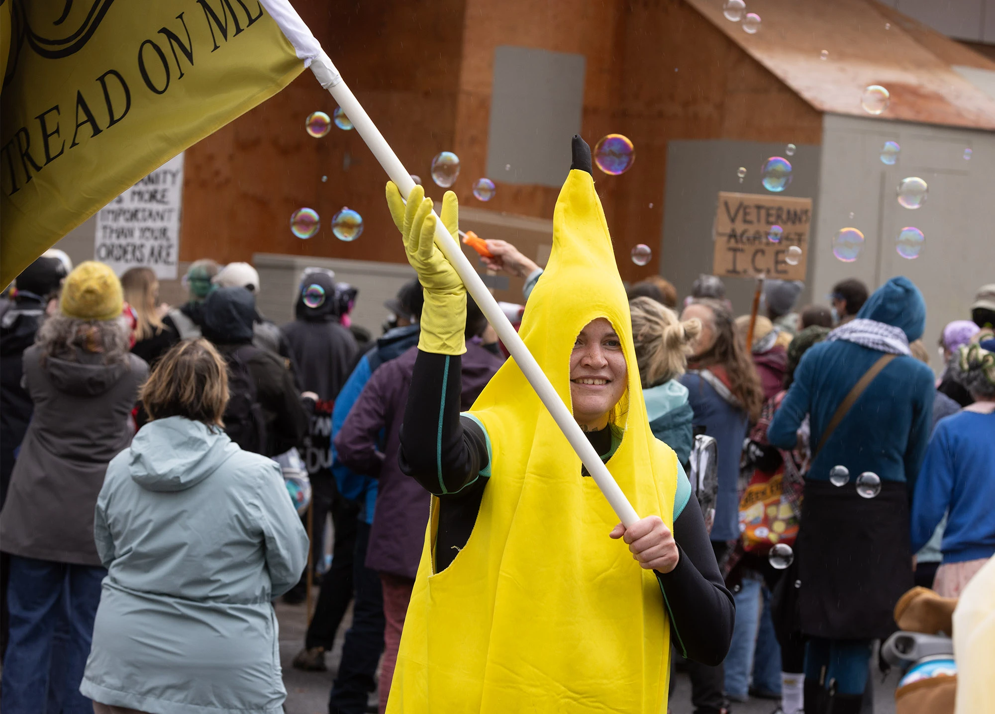 A Portland protester dressed in a bright yellow banana costume waves a 'Don't Tread On Me' flag as soap bubbles float through the air near the ICE building, blending humor and resistance during the city's anti-ICE demonstrations.