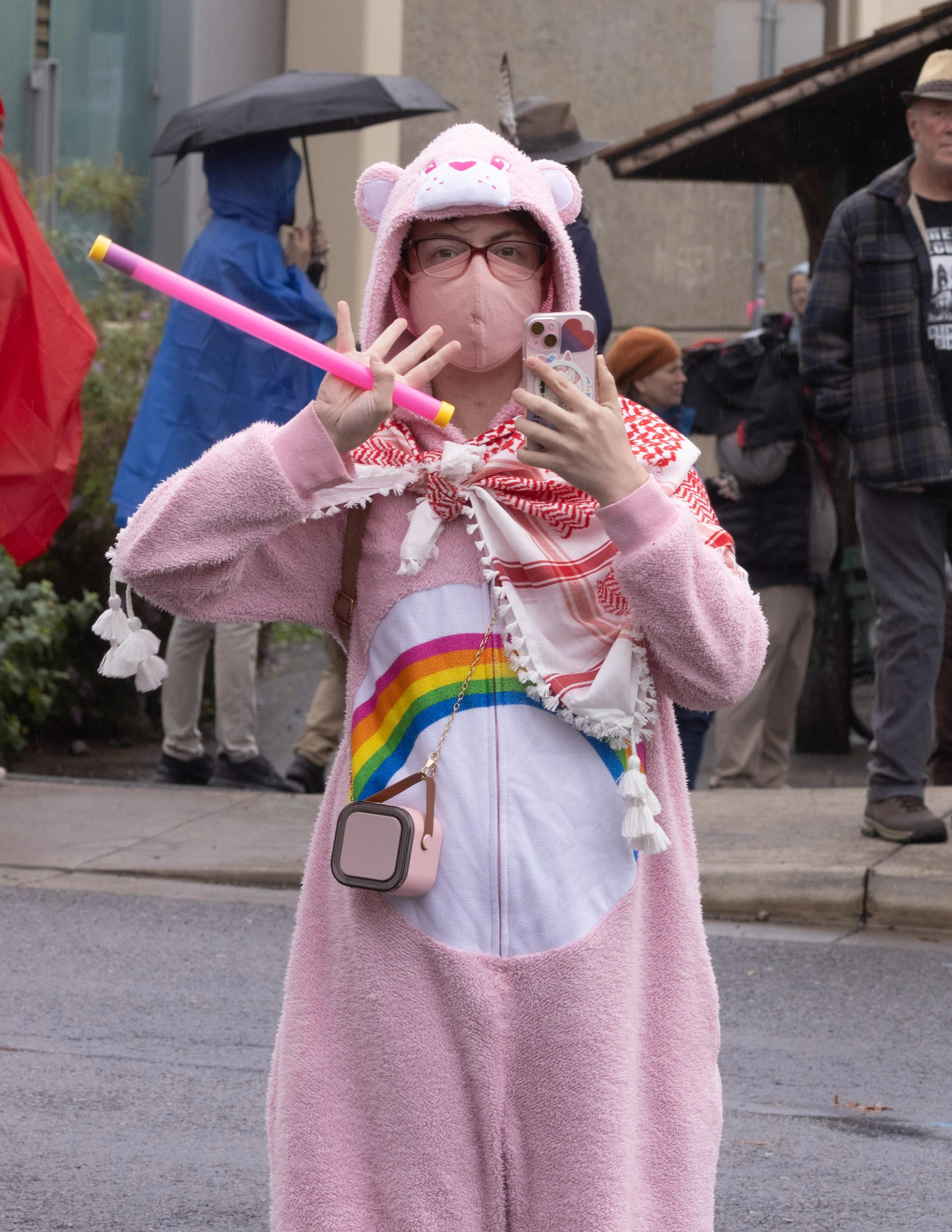 A Portland protester dressed in a pink bear costume with a rainbow emblem, matching mask, and red scarf stands in the rain holding a bright pink baton during the city's anti-ICE demonstration.