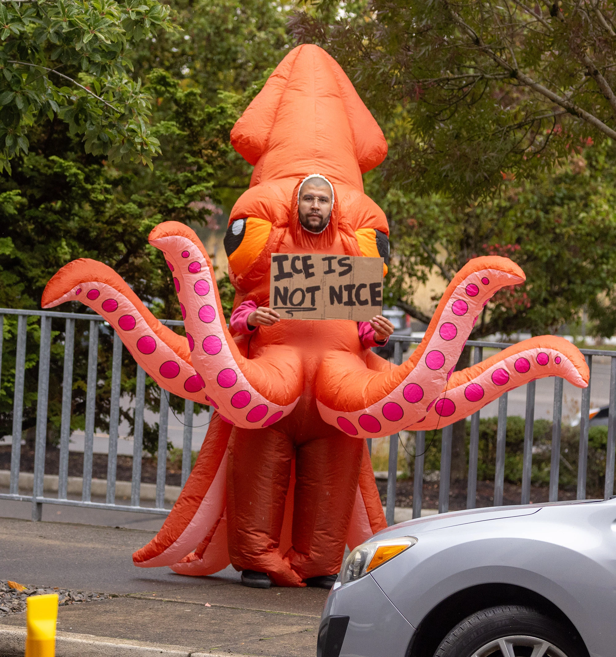 A Portland protester dressed in a bright orange inflatable squid costume holds a hand-painted cardboard sign reading 'ICE IS NOT NICE' outside the federal ICE facility, symbolizing the city's creative defiance against Trump-era immigration enforcement.