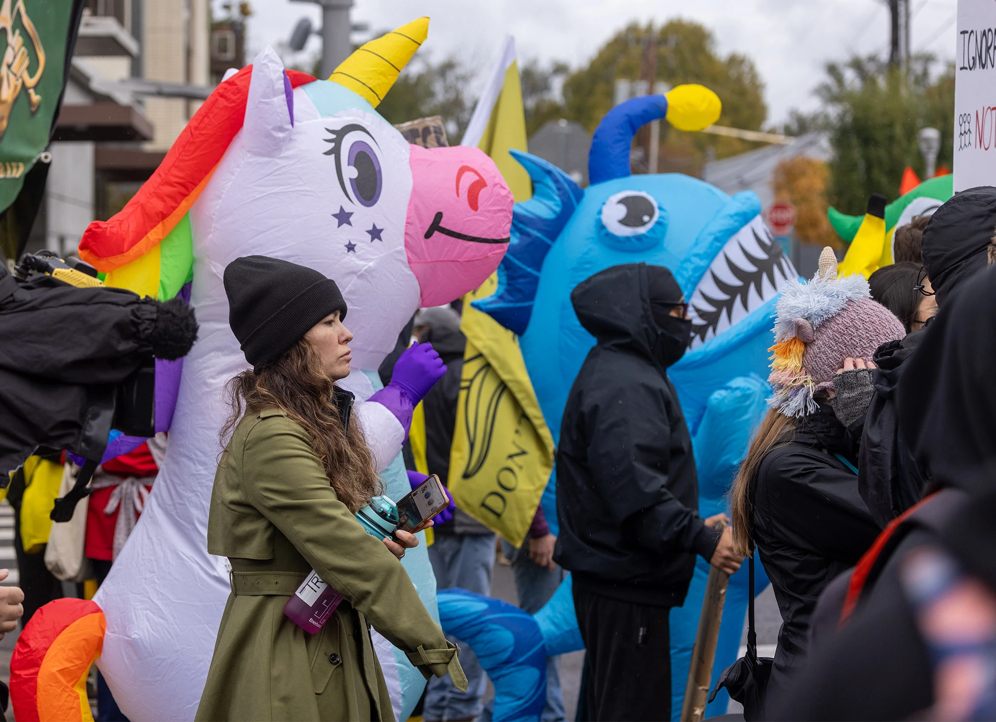 A woman in a green coat stands beside protesters dressed in colorful inflatable costumes — a rainbow unicorn and a blue shark — during Portland's anti-ICE demonstrations, blending surreal humor with defiant civic spirit.