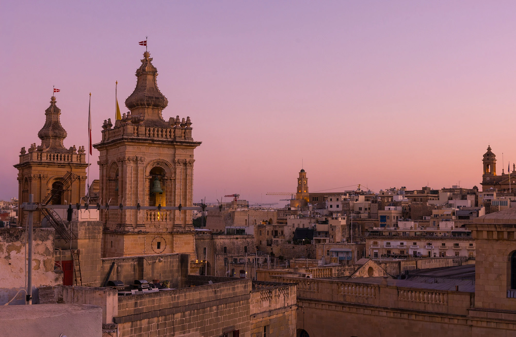 View from the rooftops of Birgu, Malta, at dusk. The ornate twin bell towers of St. Lawrence's Church rise in the foreground, their warm limestone glowing under a violet-pink sky. Beyond them, the skyline of Senglea stretches across the distance, with its own clocktower and clustered stone houses lit by the last of the evening light. Once strongholds of the Knights of St. John, these harbor cities still stand watch over the Grand Harbour.