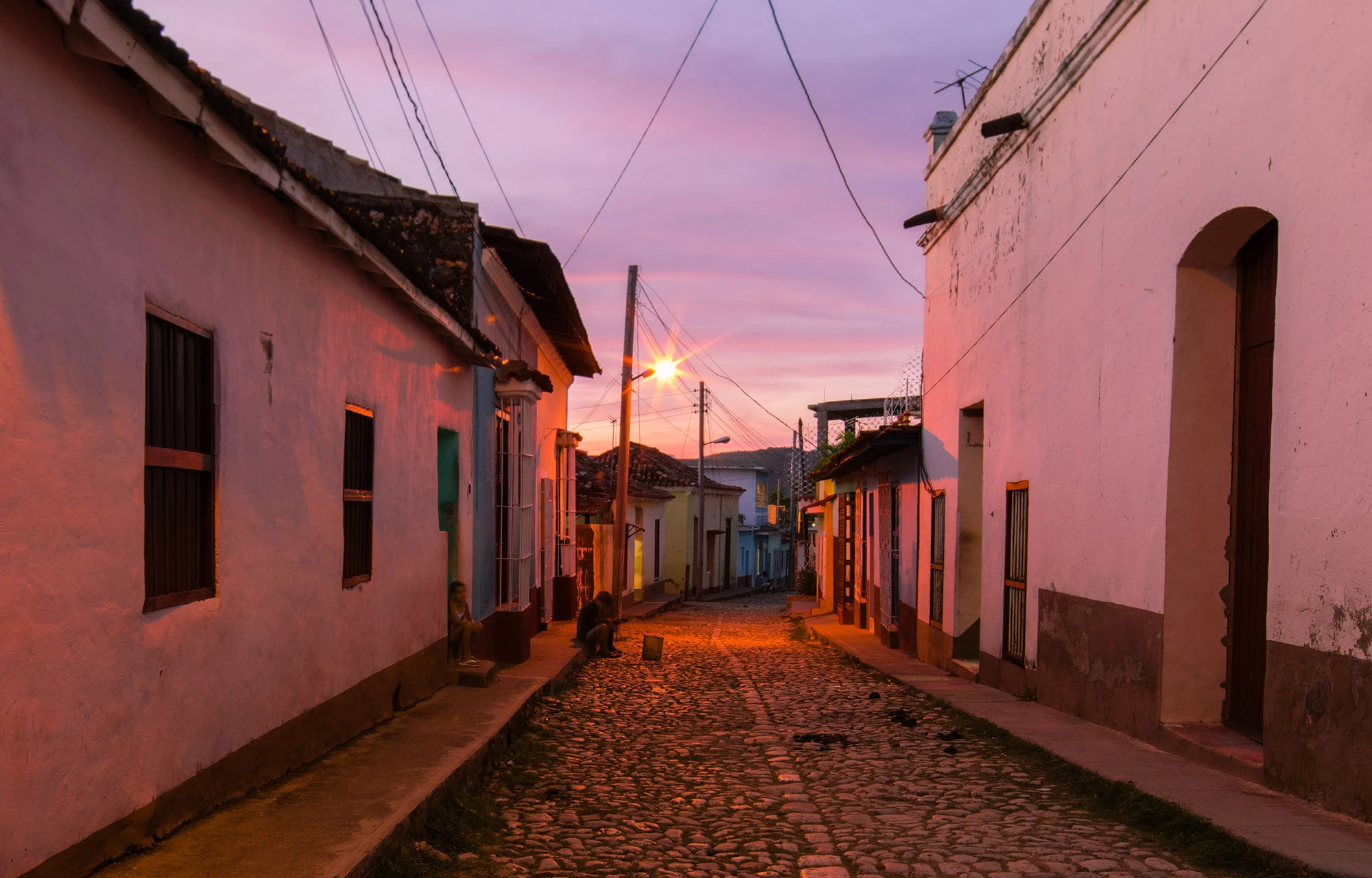 A quiet cobblestone street in Trinidad, Sancti Spíritus Province, Cuba, glows with pink and violet twilight as the last daylight fades. Streetlamps cast orange light on pastel colonial façades, while a few residents sit in doorways beneath hanging wires and tiled roofs, evoking the stillness of evening in this historic Caribbean town.