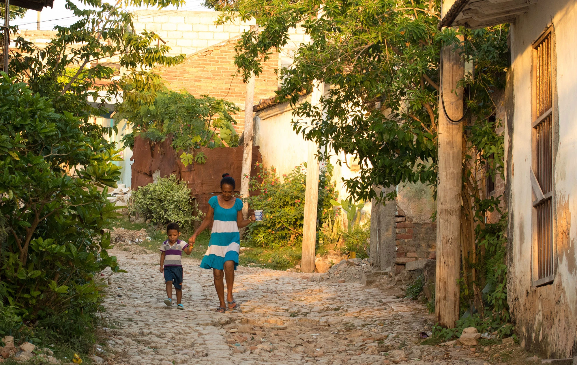 A Cuban woman and young boy walk hand in hand down a cobblestone lane in Trinidad, Sancti Spíritus Province, bathed in warm morning light. The woman carries a cup as they pass weathered walls and overgrown gardens, their striped clothing echoing the pastel hues of the old houses around them. The scene captures a quiet moment of daily life in rural Cuba.